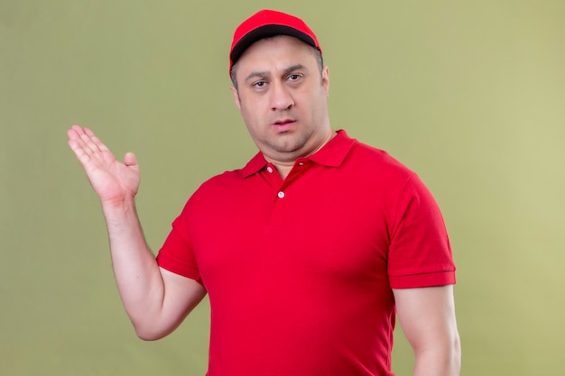 Repartidor en uniforme rojo y gorra mientras presenta con la mano con expresión seria en la cara de pie sobre un espacio verde aislado