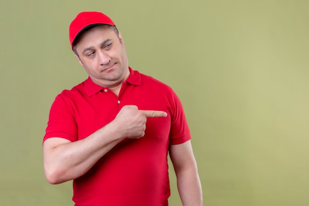 Repartidor con uniforme rojo y gorra mirando a un lado con expresión escéptica apuntando con el dedo índice hacia el lado de pie en verde aislado