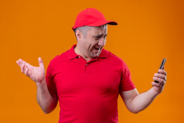 Repartidor con uniforme rojo y gorra mirando la pantalla de su teléfono inteligente sintiéndose decepcionado por la pared naranja aislada