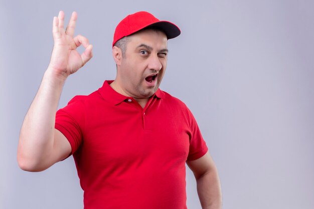 Repartidor en uniforme rojo y gorra mirando positivo y feliz sonriendo alegremente guiñando un ojo haciendo bien firmar de pie sobre un espacio en blanco aislado