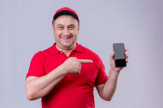 Repartidor en uniforme rojo y gorra mostrando su teléfono inteligente apuntando con el dedo índice sonriendo alegremente de pie