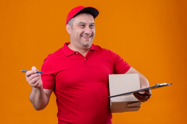 Repartidor con uniforme rojo y gorra con paquete caja y portapapeles con bolígrafo mirando a un lado con cara feliz sonriendo sobre pared naranja