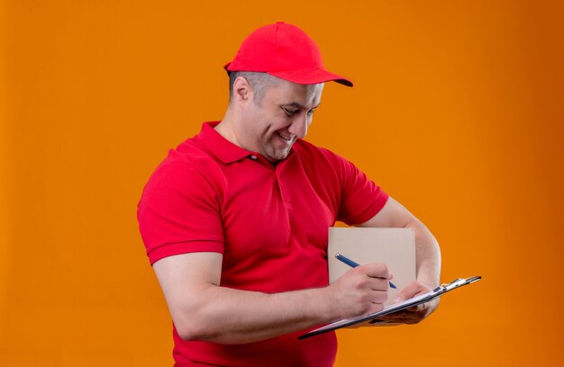 Repartidor con uniforme rojo y gorra con paquete caja y portapapeles con bolígrafo sonriendo alegremente sobre pared naranja aislada