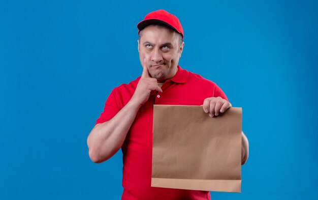 Repartidor con uniforme rojo y gorra con paquete de papel tocando su barbilla mirando hacia arriba y pensando con expresión pensativa sobre la pared azul