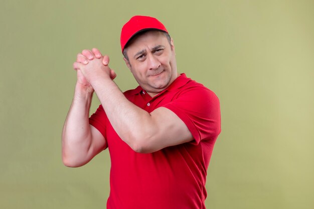 Repartidor en uniforme rojo y gorra de pie con un gesto de trabajo en equipo sonriendo confiado sobre espacio verde aislado