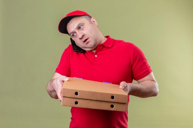 Repartidor en uniforme rojo y gorra sosteniendo cajas de pizza mientras habla por teléfono móvil con aspecto confundido de pie sobre un espacio verde aislado