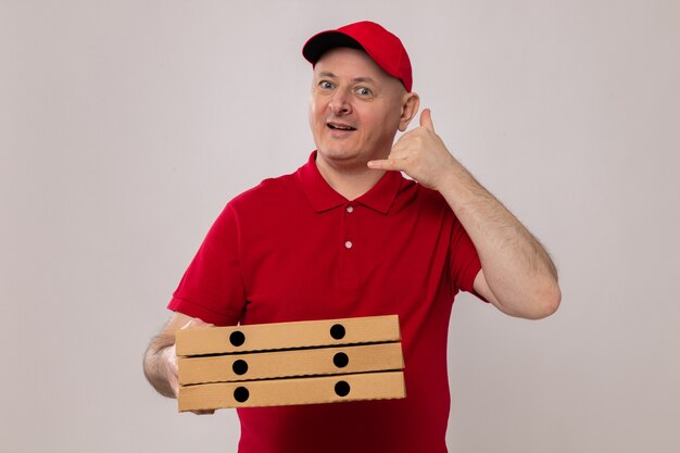 Repartidor en uniforme rojo y gorra sosteniendo cajas de pizza mirando a la cámara sonriendo feliz y positivo haciendo gesto de llamarme de pie sobre fondo blanco