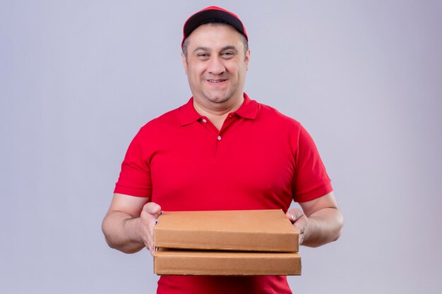 Repartidor en uniforme rojo y gorra sosteniendo cajas de pizza mirando positivo y feliz sonriendo amistoso de pie en blanco aislado