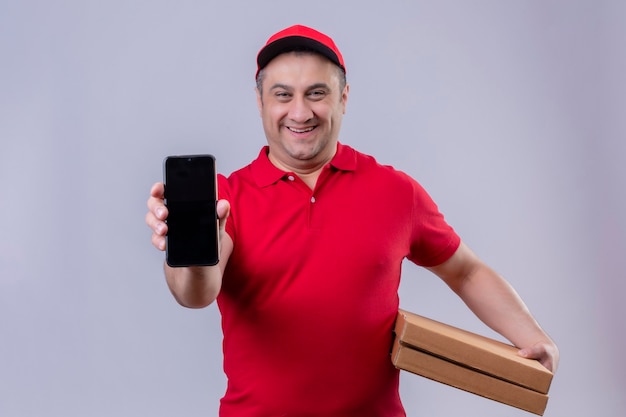 Repartidor en uniforme rojo y gorra sosteniendo cajas de pizza mostrando su smartphone sonriendo alegre con cara feliz de pie