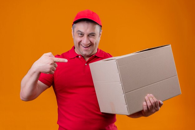 Repartidor con uniforme rojo y gorra sosteniendo una gran caja de cartón apuntando con el dedo índice sonriendo confiado sobre la pared naranja