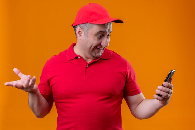 Repartidor con uniforme rojo y gorra con teléfono móvil mirando sorprendido con las manos levantadas sobre la pared naranja