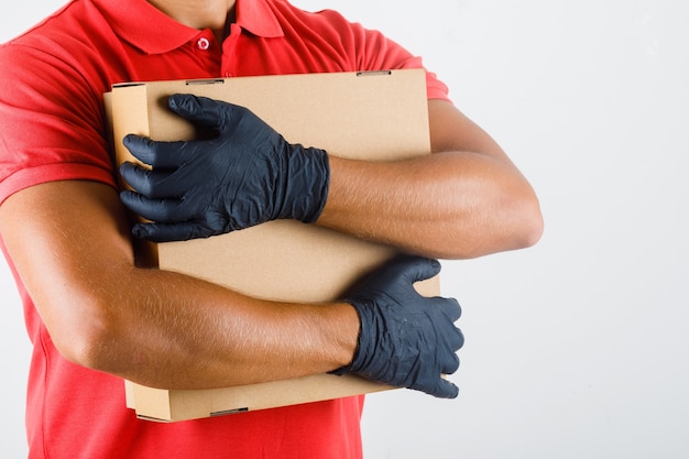 Foto gratuita repartidor en uniforme rojo, guantes médicos abrazando una caja de cartón y mirando confiado