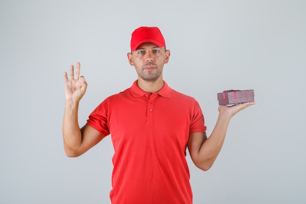Repartidor en uniforme rojo sosteniendo la caja actual y mostrando el signo de ok
