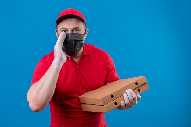 Repartidor vestido con uniforme rojo y gorra en máscara protectora facial sosteniendo cajas de pizza de pie con una mano cerca de la boca contando un secreto sobre un espacio azul aislado