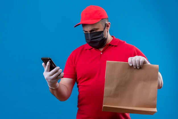 Repartidor vestido con uniforme rojo y gorra en máscara protectora facial sosteniendo el paquete de papel y mirando la pantalla de su teléfono móvil con cara seria de pie sobre el espacio azul