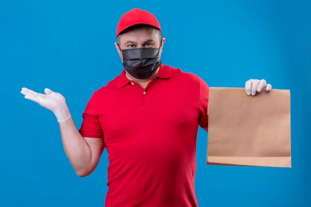 Foto gratuita repartidor vestido con uniforme rojo y gorra en máscara protectora facial sosteniendo el paquete de papel que presenta con el brazo oh mano de pie sobre el espacio azul