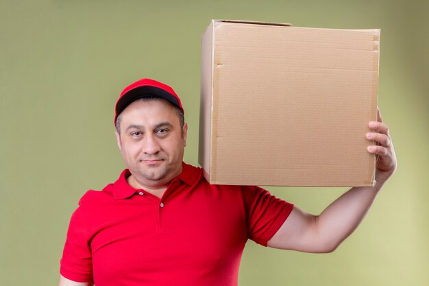 Repartidor vestido con uniforme rojo y gorra sosteniendo una caja de cartón en el hombro mirando confiado con una sonrisa en la cara de pie sobre un espacio verde aislado