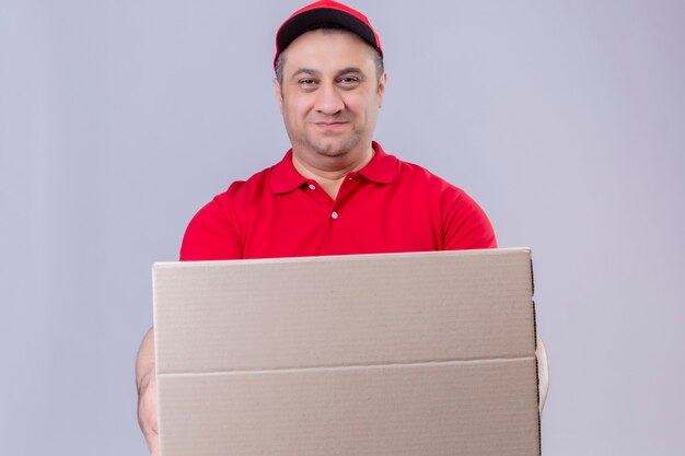 Repartidor vestido con uniforme rojo y gorra sosteniendo una caja de cartón mirando confiado sonriendo de pie sobre un espacio en blanco aislado