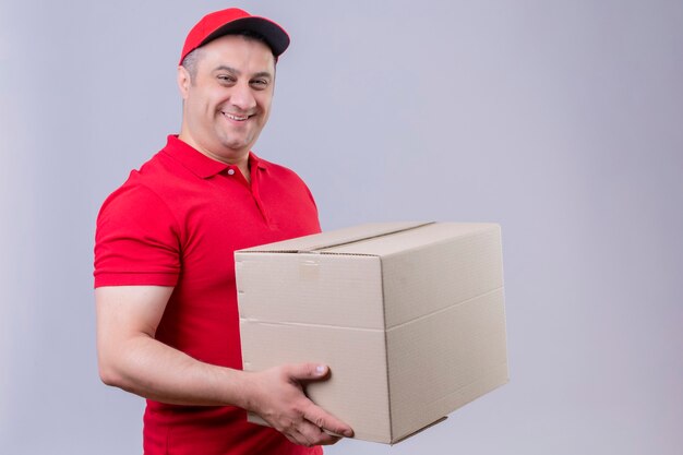 Repartidor vestido con uniforme rojo y gorra sosteniendo una caja de cartón mirando confiado con una sonrisa en la cara de pie sobre un espacio en blanco aislado