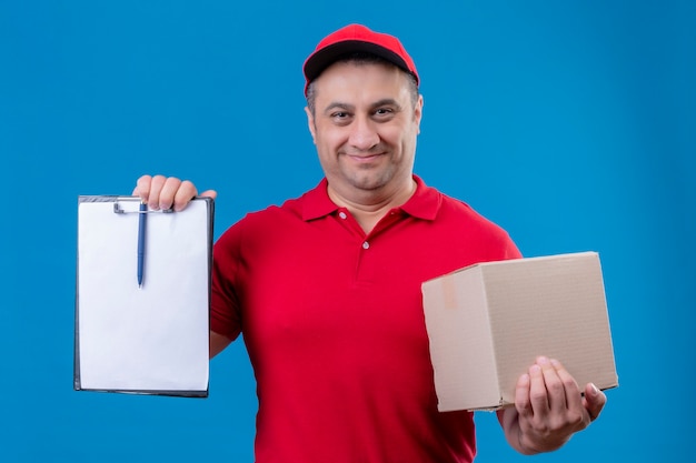 Repartidor vestido con uniforme rojo y gorra sosteniendo una caja de cartón que muestra el portapapeles con espacios en blanco mirando confiado sonriendo de pie sobre el espacio azul