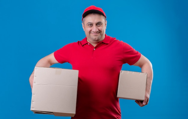 Foto gratuita repartidor vestido con uniforme rojo y gorra sosteniendo cajas de cartón mirando confiado sonriendo parado sobre espacio azul aislado