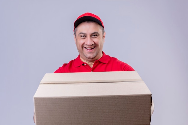 Repartidor vestido con uniforme rojo y gorra sosteniendo una gran caja de cartón sonriendo amable con cara feliz de pie sobre un espacio en blanco aislado