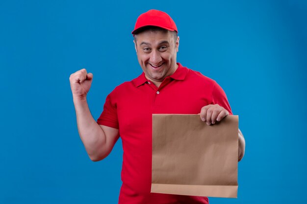 Repartidor vestido con uniforme rojo y gorra sosteniendo el paquete de papel cara sonriente levantando el puño después de una victoria de pie sobre el espacio azul aislado