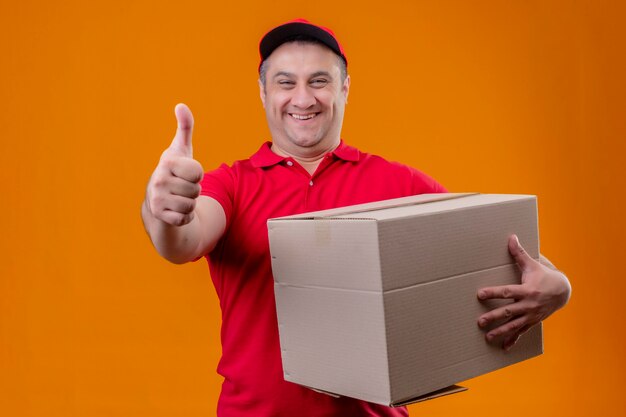 Repartidor vestido con uniforme rojo y gorra sosteniendo el paquete de papel salió y feliz levantando el puño después de una victoria de pie sobre el espacio azul aislado sosteniendo el paquete de caja mirando vino