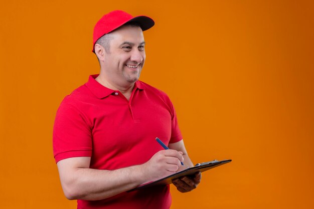 Repartidor vestido con uniforme rojo y gorra sosteniendo portapapeles mirando a un lado positivo y feliz escribiendo algo sonriente de pie