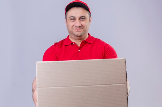 Repartidor vistiendo uniforme rojo y gorra con caja de cartón mirando confiado sonriendo sobre pared blanca aislada