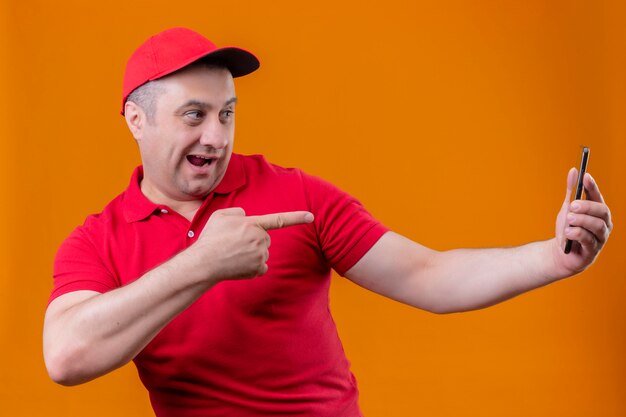 Repartidor vistiendo uniforme rojo y gorra mirando alegre celebración smartphone y señalando con el dedo índice sonriendo sobre la pared naranja