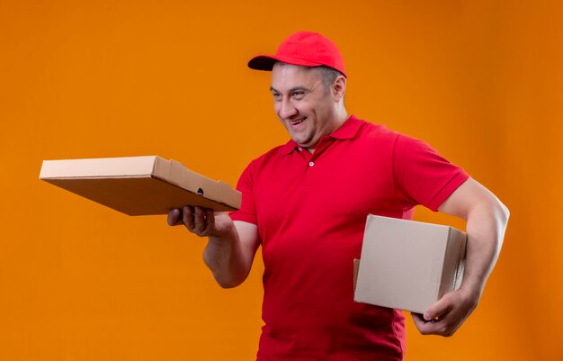 Repartidor vistiendo uniforme rojo y tapa con paquete de caja dando pizza a un cliente sonriendo alegre sobre pared naranja aislada