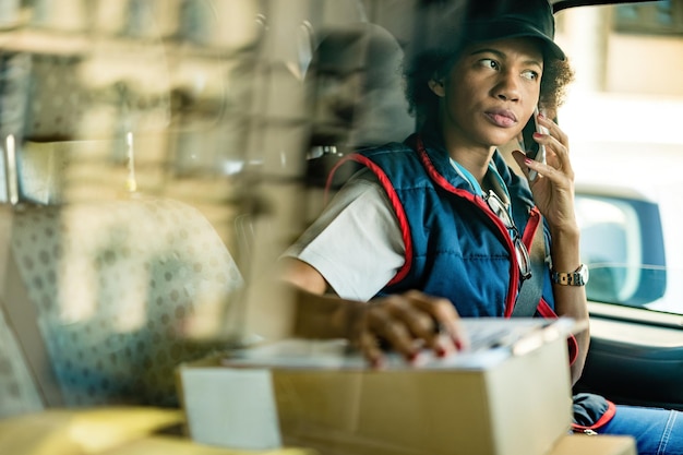 Repartidora afroamericana hablando con el cliente por teléfono móvil mientras está sentada en una mini furgoneta La vista es a través de la ventana