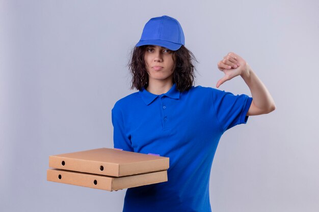 Repartidora disgustada en uniforme azul y gorra con cajas de pizza mostrando los pulgares hacia abajo sobre la pared blanca