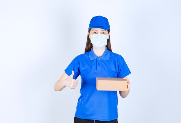 Repartidora joven en máscara médica sosteniendo la caja de cartón y dando los pulgares para arriba.