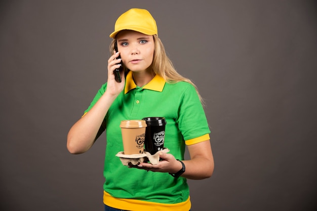 Repartidora joven con tazas de café hablando por teléfono inteligente.