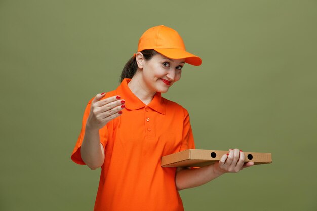 Repartidora de mediana edad complacida con uniforme y gorra sosteniendo un paquete de pizza mirando a la cámara haciendo un gesto de venir aquí aislado en un fondo verde oliva