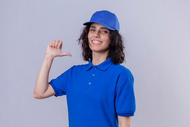 Repartidora en uniforme azul y gorra apuntando con el pulgar sobre sí misma sonriendo confiada autosatisfecha de pie sobre un espacio en blanco aislado