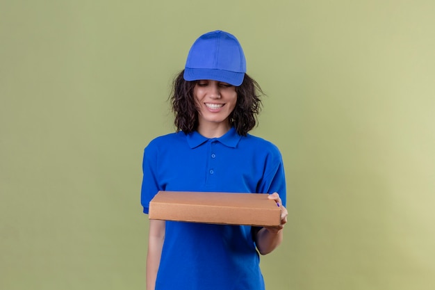 Foto gratuita repartidora en uniforme azul y gorra con caja de pizza sonriendo alegre mirando cuadro de pie en color oliva aislado