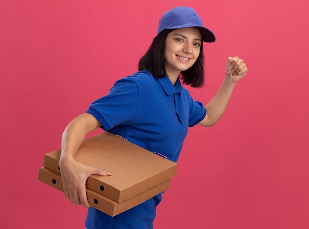 Repartidora en uniforme azul y gorra corriendo para entregar cajas de pizza para el cliente sobre una pared rosa