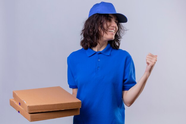 Repartidora en uniforme azul y gorra sosteniendo cajas de pizza apuntando hacia atrás con la mano y el pulgar sonriendo alegremente de pie en blanco