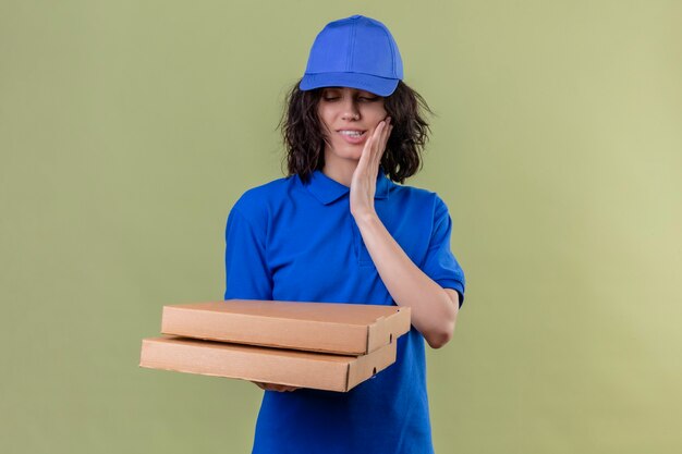 Repartidora en uniforme azul y gorra sosteniendo cajas de pizza mordiéndose el labio con expresión hambrienta sonriendo de pie sobre el espacio de color oliva aislado