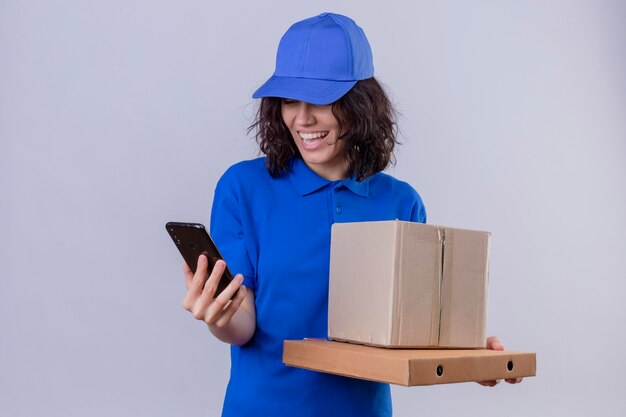 Foto gratuita repartidora en uniforme azul y gorra sosteniendo cajas de pizza y paquete de caja mirando la pantalla del teléfono móvil sonriendo con cara feliz de pie en blanco