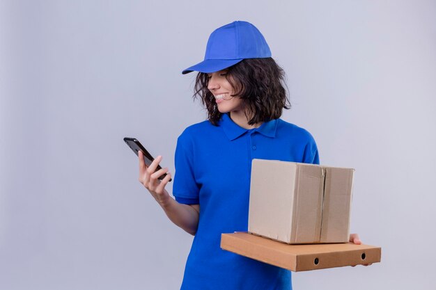 Repartidora en uniforme azul y gorra sosteniendo cajas de pizza y paquete de caja mirando la pantalla del teléfono móvil sonriendo con cara feliz de pie