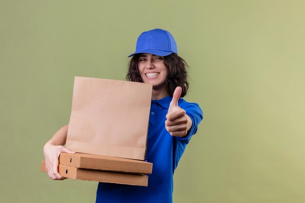 Repartidora en uniforme azul y gorra sosteniendo cajas de pizza y paquete de papel sonriendo alegremente mostrando los pulgares para arriba sobre un espacio verde aislado