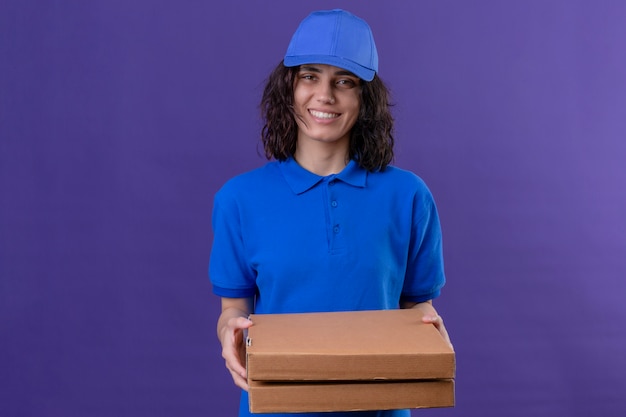 Repartidora en uniforme azul y gorra sosteniendo cajas de pizza positivo y feliz sonriendo amable de pie en púrpura aislado