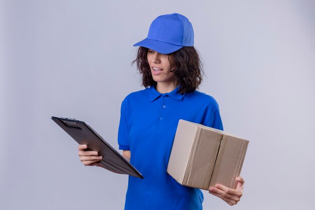 Repartidora en uniforme azul y gorra sosteniendo el paquete de caja y mirando el portapapeles con una sonrisa en la cara de pie