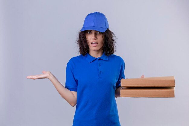 Repartidora en uniforme azul sosteniendo cajas de pizza con aspecto incierto y confundido, sin respuesta extendiendo palmas de pie en blanco