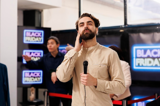 Foto gratuita reportero de televisión transmitiendo en vivo hablando de las líneas del black friday en el centro comercial local, sosteniendo el micrófono y mirando a la cámara. joven corresponsal cubriendo noticias sobre la temporada de ventas en el comercio minorista