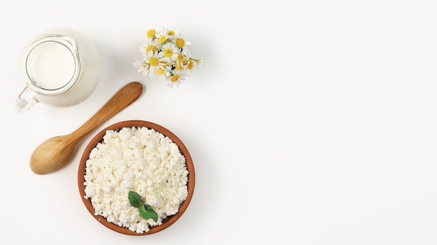 Requesón granulado en un cuenco de barro, junto a una cuchara de madera y una jarra de leche. Diseño sobre fondo blanco brillante con espacio de copia. Requesón blando, comida sana natural, comida dietética completa